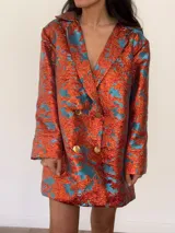 Blazer vestido de corte oversize con doble botonadura dorada, cuello con solapa y mangas largas, confeccionado en tejido jacquard texturado con acabado brillante en tonos naranja y celeste.