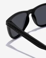 Lentes de sol unisex Hawkers modelo One Raw Carbono, con montura cuadrada negra con textura de fibra de carbono y lentes polarizadas oscuras.