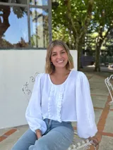 Blusa blanca de algodón con textura arrugada, cuello redondo, mangas largas abullonadas con puños elásticos y cierre frontal con botones y volantes.