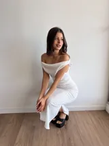 Conjunto de dos piezas en color blanco, compuesto por un top con escote bote y hombros descubiertos, y una falda midi con tajo lateral. Ambas prendas están confeccionadas en tela bambula elastizada con textura arrugada.