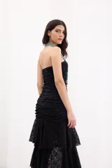 Vestido largo negro strapless de encaje con frunces en el torso, falda asimétrica en capas con volados y breteles removibles.