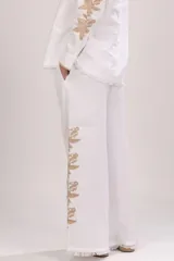 Pantalón blanco de lino, de pierna recta y corte holgado, con cintura elastizada, cordón ajustable con pompones y detalle de bordado lateral en tonos neutros. Presenta terminación desflecada en el bajo.