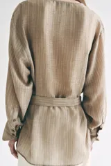 Camisa de cuello fluido y manga larga con puños anchos, de corte recto y largo medio, confeccionada en tejido rústico en tono beige con sutil textura jaspeada. Cuenta con un cinturón ajustable del mismo género para entallar la cintura.