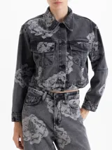 Chaqueta cropped de denim con lavado gris y estampado floral en contraste. Presenta cuello clásico, mangas largas, bolsillos frontales con tapa y cierre mediante broches metálicos.