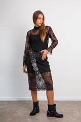 Conjunto de dos piezas compuesto por blusa de encaje semitransparente con cuello alto y mangas largas, y falda midi a juego. Ambas prendas incluyen visos interiores a tono. El diseño presenta un encaje floral delicado en color negro.