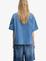 Camisa de denim de algodón celeste, de corte holgado y mangas cortas, con cierre frontal de botones metálicos y un bolsillo aplicado en el pecho con etiqueta de la marca.