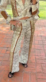 Pantalón acampanado de tiro medio, confeccionado en tela brocada con estampado floral en tonos beige y dorado, con franjas laterales de lentejuelas doradas y ribetes de cuero marrón.