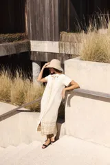 Vestido largo de lino en color beige, con cuello camisero, botones frontales y mangas cortas. Presenta bordados decorativos en contraste a lo largo de la silueta y en el ruedo.