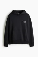Canguro de corte oversized en mezcla de algodón, color negro, con capucha de doble capa, hombros caídos y bolsillo frontal tipo canguro. Presenta un estampado tipográfico pequeño en el pecho y puños y bajo acanalados.