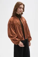 Chaqueta tipo bomber color tostado, confeccionada en material efecto gamuza. Presenta diseño con frunces en el pecho y espalda, cuello mao, cierre frontal completo y mangas largas con puños elásticos.