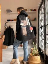 Campera con capucha de textura tipo corderito en color negro, con un diseño de collage geométrico en la espalda que combina parches en tonos marrón, negro, blanco y rojo. Presenta un corte holgado y diseño artesanal único.