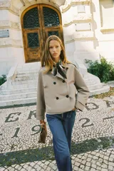 Chaqueta corta estilo trench color marrón oscuro, de corte cruzado con doble botonadura, cuello solapa y hombreras. Presenta mangas largas con puños anchos y bolsillos delanteros de vivo.