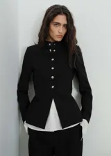 Chaqueta entallada de color negro con cuello mao y cierre frontal de botones metálicos efecto joya. Presenta diseño de largo medio, manga larga y forro interior.