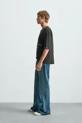 Camiseta de punto con corte boxy fit, cuello redondo y manga corta. Color negro con efecto lavado y terminaciones irregulares. Presenta un estampado frontal con texto y un gráfico circular en tonos blanco y rojo.