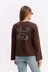Remera de manga larga color beige con silueta relajada y mangas amplias. Presenta una estampa en la espalda con la frase 'Be kind to your mind' acompañada de ilustraciones de un cupido, flores y lazos en tono bordó.