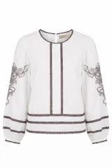 Blusa blanca de manga larga con detalles de bordados florales en tono grisáceo y bandas decorativas en el frente y mangas. Cuenta con cuello redondo y puños elásticos.