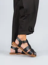 Sandalias planas de cuero negro estilo pescador, con tiras anchas entrelazadas en el empeine, suela marrón y tira ajustable con hebilla metálica en el tobillo.