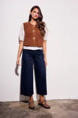 Chaleco tejido color beige con cuello redondo, sin mangas, con cierre frontal de cuatro botones dorados y dos bolsillos frontales.