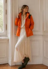 Piloto impermeable color naranja con capucha integrada, cintura regulable con cordón y tranca, mangas con volumen y puños elastizados. Presenta detalles decorativos en color verde oliva.