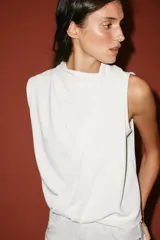 Top blanco de manga sisa con cuello redondo y diseño drapeado cruzado en la parte frontal, con bajo asimétrico y cierre en la espalda con abertura y botones.