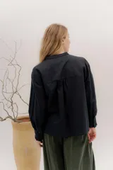 Camisa de algodón blanca con cuello a la base, cierre frontal con botones y mangas largas con puños delicados. Presenta volados verticales y detalles de cintas de terciopelo en contraste a lo largo del frente.