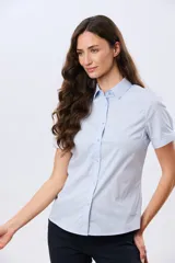 Camisa celeste rayada de manga corta, con cuello camisero y cierre frontal con botones. Presenta un corte entallado y un pequeño detalle de placa metálica en el borde inferior.
