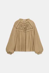 Camisa blanca de manga larga con cuello redondo y diseño abullonado, con detalle de bordados y encaje en la parte frontal y cierre de botón en la espalda.