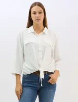 Camisa blanca de rayón con textura sutil, cuello con solapa, cierre frontal con botones y bolsillos simulados en el pecho. Presenta mangas 3/4 regulables con presilla y un diseño con nudo en el ruedo frontal.