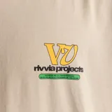Remera color beige con estampado pequeño en el pecho con el logo de la marca Rivvia en amarillo y verde.