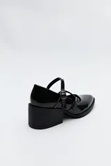 Zueco estilo Mary Jane color negro charol, con doble tira y hebillas metálicas. Presenta punta cuadrada y taco ancho de altura media.