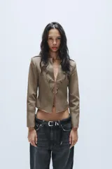 Blazer corto de estilo militar color tostado, con cuello redondo, manga larga y cierre frontal con ganchos metálicos. Presenta detalle de botones decorativos dorados en el frente y en los hombros, con costuras marcadas y forro interior a contraste.