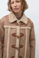 Abrigo de estilo duffle coat en simil gamuza color tostado, con interior y bordes de piel sintética al tono. Presenta cierre frontal mediante alamares de cuero sintético y madera, cuello solapa y mangas largas.