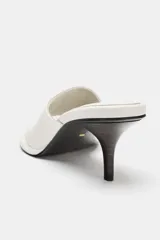 Sandalia tipo mule de tacón alto fino de madera, confeccionada en piel color blanco marfil. Presenta una tira asimétrica con separador para el dedo y punta redonda.