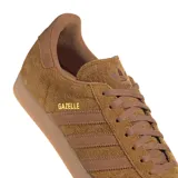 Championes adidas modelo Gazelle, confeccionados en gamuza color marrón con las icónicas tres tiras laterales en tono a juego. Presentan suela de goma color caramelo y el nombre del modelo grabado en dorado en el lateral.