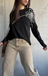 Remera de manga larga color negro, de corte recto, con aplique decorativo en relieve con forma de flor grande en el hombro confeccionado con cordón trenzado en blanco y negro.