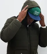 Campera puffer de hombre color verde militar, con capucha integrada, cierre frontal por cremallera y bolsillos cargo frontales. Presenta un diseño acolchado con acabado mate y ajuste en las mangas.