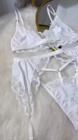 Conjunto de lencería de cuatro piezas en color blanco, confeccionado en lycra y encaje. Incluye un bralette triangular, una tanga regulable y un portaligas con ligas integradas, todo decorado con detalles de encaje en los bordes.
