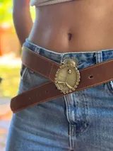 Cinturón de cuero marrón grueso con pespuntes, ajustado a la cintura. La hebilla es redonda, de metal dorado o bronce, con el Escudo Nacional Argentino en relieve rodeado por una corona de laureles y un sol en la parte superior.