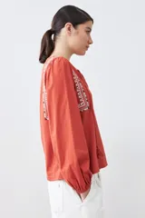 Remera blanca de manga larga con detalles bordados en tonos azules y verdes en el pecho y hombros. Presenta un escote en V con cordones ajustables terminados en borlas y puños elásticos.