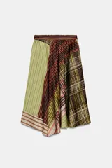 Falda midi de tiro alto con bajo asimétrico. Presenta un diseño patchwork con bloques de diferentes estampados, incluyendo rayas verticales en verde lima, rayas diagonales en marrón oscuro y naranja, y un estampado a cuadros en tonos verdes y marrones.