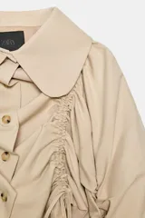 Chaqueta corta color beige, confeccionada en gabardina, con cuello solapa redondeado, mangas largas con frunces y detalle de frunces con lazada en el delantero, cerrando con botones.