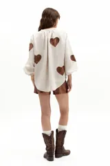Camisa oversize de lino color beige, con cuello mao, cierre frontal con botones de nácar y mangas abullonadas con puño elástico. Presenta apliques de corazones en material metalizado tono bronce.