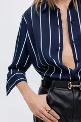 Camisa de manga larga con cuello clásico, cierre frontal abotonado y puños con botón. Presenta un diseño de rayas verticales blancas sobre fondo azul marino, con bajo redondeado.