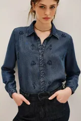 Camisa de denim azul oscuro con cuello clásico, cierre frontal de botones y mangas largas. Presenta un delicado bordado de motivos florales y vegetales realizado con cuentas y canutillos en tono azul marino.