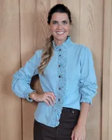 Camisa de denim celeste con cuello alto y volados frontales a lo largo de la botonadura. Presenta mangas largas con puños elásticos y terminación con volados.