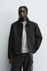 Cazadora de algodón color negro lavado, de corte relajado (relaxed fit), con cuello solapa a contraste, cierre frontal de cremallera, bolsillos canguro en la cadera y puños con botón a presión.