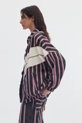 Campera de denim oversize con cierre frontal, cuello alto y bolsillos delanteros. Presenta un diseño a rayas verticales en tonos negros y rosados, cruzado por una franja ancha horizontal de color blanco con forma de zigzag.