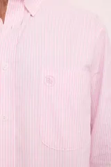 Camisa de corte clásico confeccionada en algodón, con diseño de rayas verticales finas en color rosa sobre fondo blanco. Presenta cuello abotonado, cierre frontal con botones, mangas largas con puños abotonados y un bolsillo en el pecho con el logo de la marca bordado.