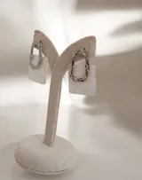 Par de aros colgantes con diseño geométrico compuesto por una pieza superior metálica de forma irregular y una pieza inferior cuadrada de resina con efecto nacarado.