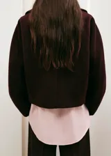 Chaqueta corta bordó confeccionada en mezcla de lana, con cuello amplio solapado, cierre frontal de botones oscuros y bolsillos ribeteados en los laterales. Presenta mangas anchas y caídas de corte relajado.